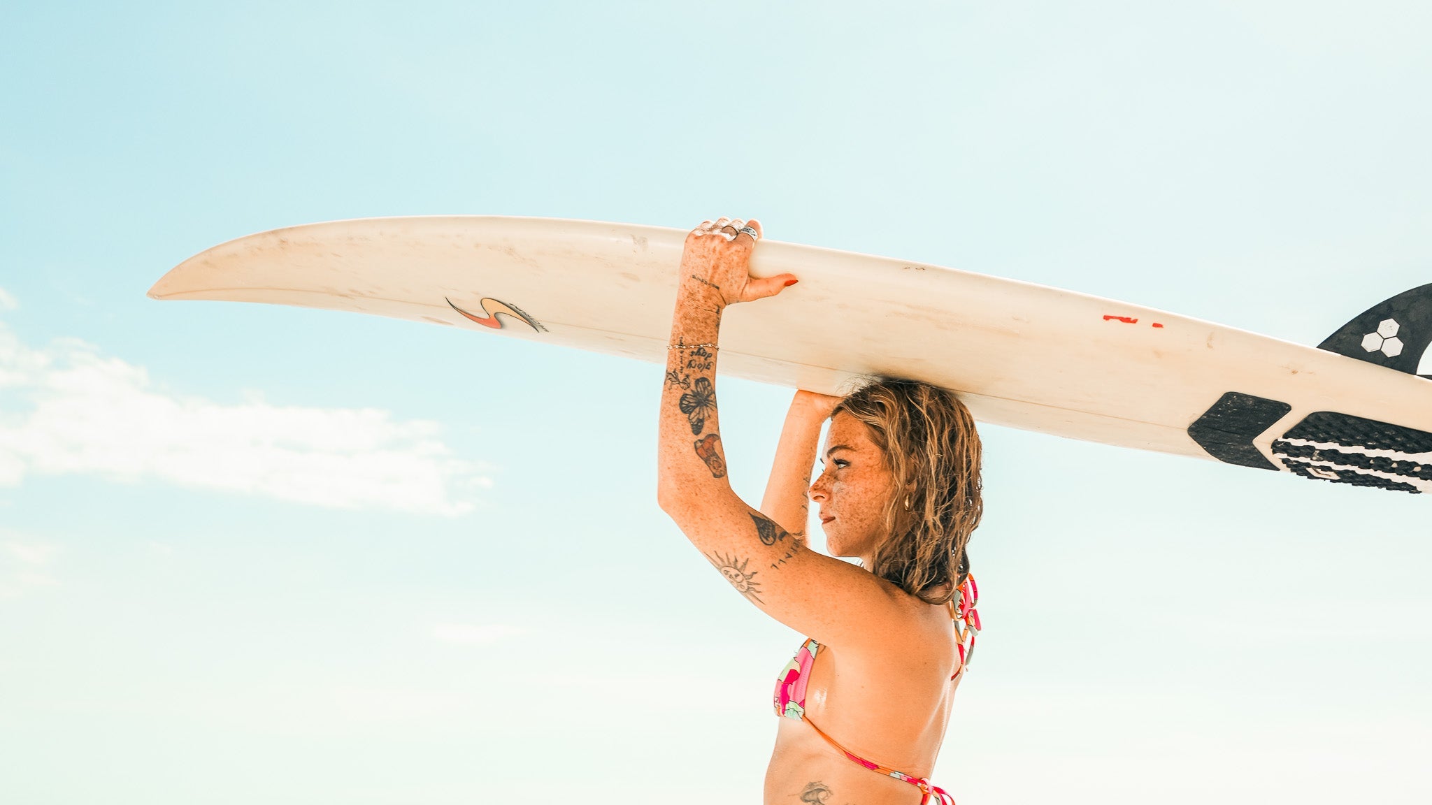 Woman in a bikini holding a surfboard on a beach with clear blue sky and green water.